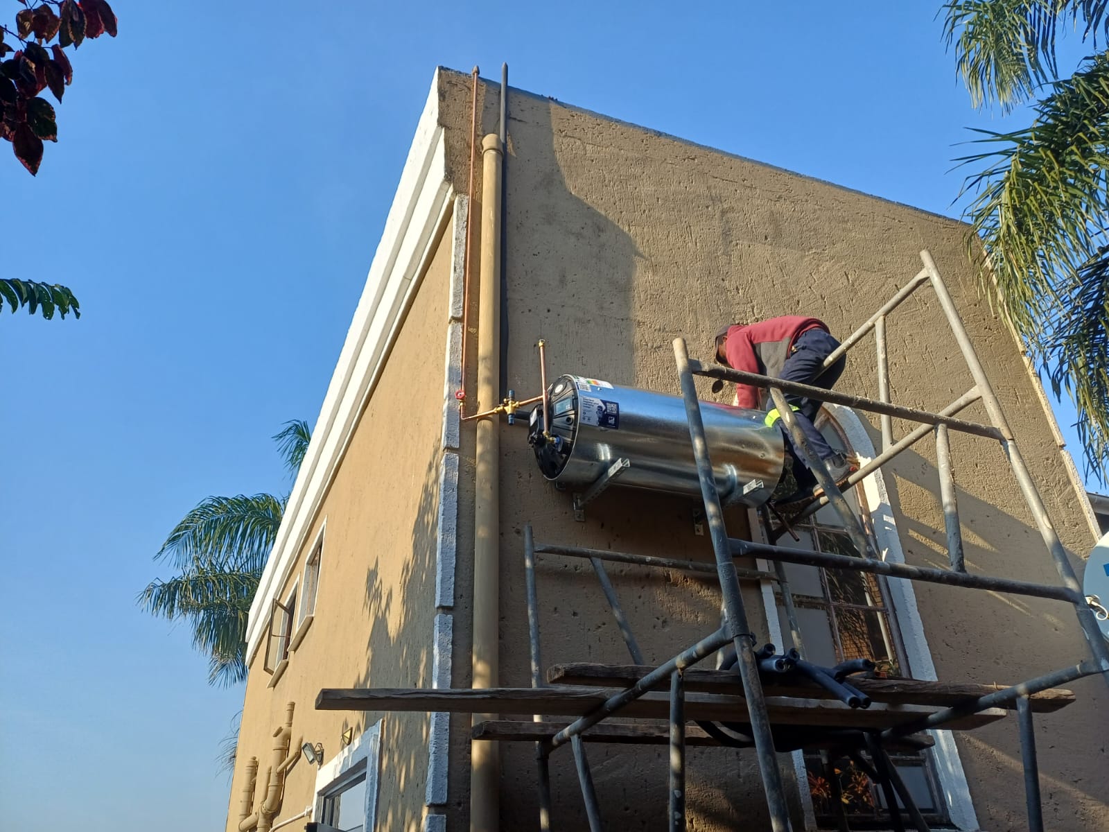 Technician installing a new geyser high on an exterior wall from scaffolding